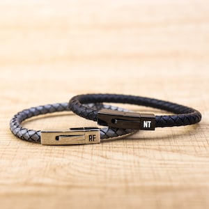 May include: Two leather bracelets with silver clasps. The top bracelet is black and has the initials "NT" on the clasp. The bottom bracelet is grey and has the initials "RF" on the clasp.