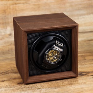 K&ouml;nnte beinhalten: Ein Uhrenbeweger aus Holz mit schwarzem Innenraum und einer Uhr darin. Der Uhrenbeweger steht auf einer Holzoberfl&auml;che.