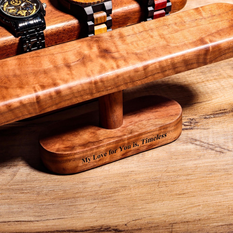 May include: Wooden watch stand with a brown finish. The stand has a rectangular base and a curved top. The stand is engraved with the text "My Love for You is, Timeless".