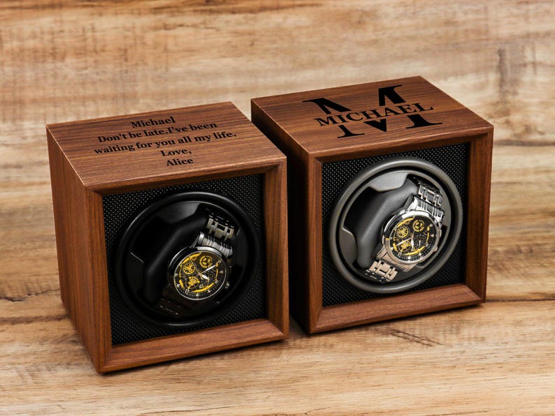 K&ouml;nnte beinhalten: Zwei Uhrenbeweger aus Holz mit schwarzem Innenraum und einer Uhr darin. Ein Beweger hat den Text "Michael Don't be late. I've been waiting for you all my life. Love, Alice" auf der Oberseite eingraviert. Der andere Beweger hat den Text "MICHAEL IV" auf der Oberseite eingraviert.