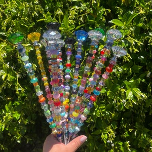 Op de afbeelding: Een verzameling kleurrijke, met kralen versierde tuinpalen met diverse juweelkleurige glazen toppers. De palen hebben een verscheidenheid aan kralen in verschillende vormen, maten en kleuren, waaronder rood, oranje, geel, groen, blauw en helder. De toppers zijn facetgeslepen.