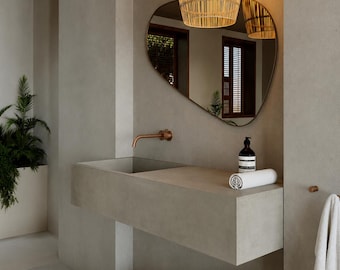 May include: A bathroom vanity featuring a rectangular concrete sink basin with a copper wall-mounted faucet, an irregular organic-shaped mirror, a woven rattan pendant light, a white rolled towel, a dark soap bottle, and a woven basket.