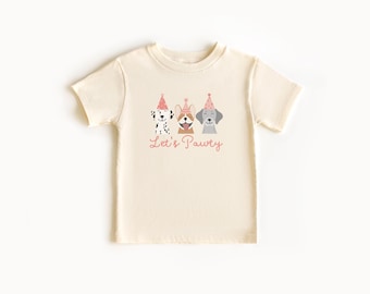 Puede incluir: Una camiseta de manga corta de color crema con un gráfico de tres perros que llevan gorros de fiesta rosas. Debajo de los perros, el texto Let's Pawty está impreso en una fuente cursiva rosa. El diseño está centrado en el pecho.
