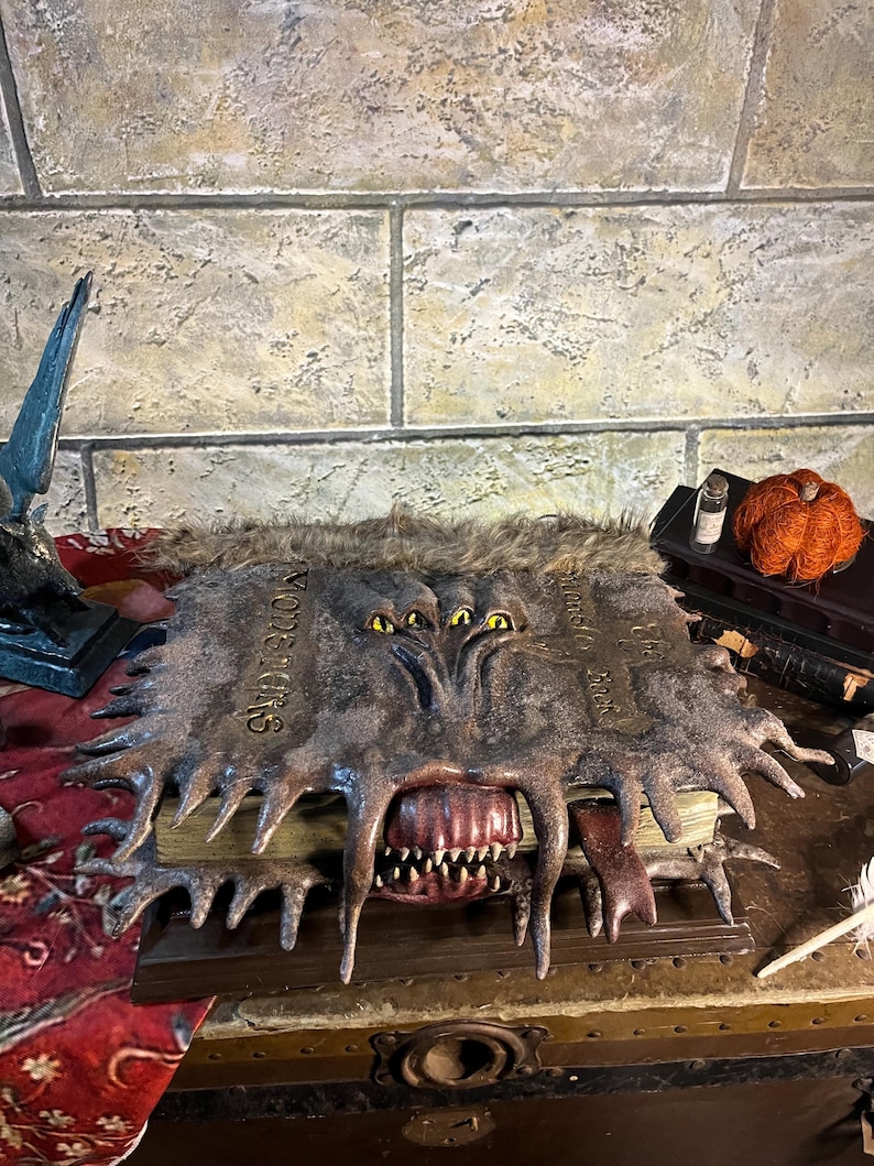 Op de afbeelding: Een gedetailleerd, open boek met een monsterlijk ogende kaft. De kaft is donkergrijs met gele ogen, scherpe tanden en een lange tong. Het boek is open en onthult oude pagina's.