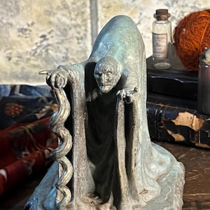 May include: A bronze-toned statue of a cloaked figure holding a serpent. The figure has a gaunt face and is standing on a base. A small glass bottle with a cork stopper and a ball of orange yarn are in the background.