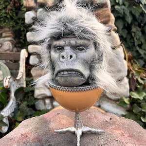 Puede incluir: Una escultura de cerámica de una cabeza de mono con pelo blanco, sentada dentro de una taza de color marrón en forma de huevo con un borde plateado. La taza está sobre un soporte plateado de tres puntas.