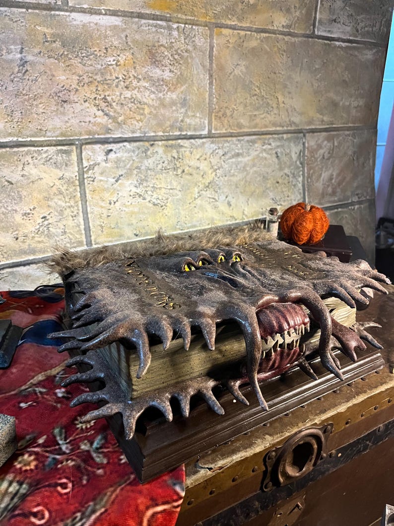 Op de afbeelding: Een gedetailleerd, open boek met een monsterlijk ogende kaft. De kaft is bruin met gekartelde randen en gele ogen. De open pagina's onthullen een rode mond met witte tanden. Een kleine oranje pompoen staat in de buurt.