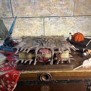 Op de afbeelding: Een gedetailleerd, open boek met een monsterlijk gezicht dat in de kaft is gesneden. De kaft is bruin en harig, met meerdere gele ogen en scherpe tanden. Het boek staat op een houten kist, met andere decoratieve items.