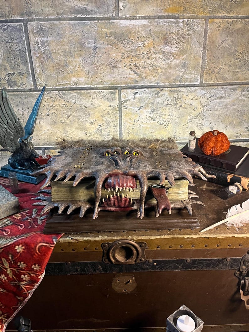 Op de afbeelding: Een gedetailleerd, open boek met een monsterlijk gezicht dat in de kaft is gesneden. De kaft is bruin en harig, met meerdere gele ogen en scherpe tanden. Het boek staat op een houten kist, met andere decoratieve items.