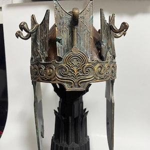 Peut inclure: Une couronne dorée et verte avec une base noire. La couronne présente un design détaillé avec des serpents sur les côtés et un design ailé sur le dessus.