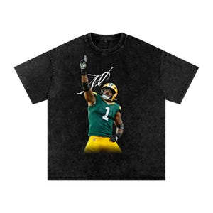 May include: Black t-shirt featuring a football player in a green and yellow uniform, raising his arm with one finger pointing up. The shirt has a distressed look and a white signature above the player.