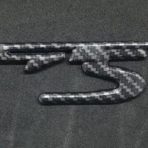 Op de afbeelding: Een zwart en grijs carbonfiber embleem met een gestileerd "S"-ontwerp.