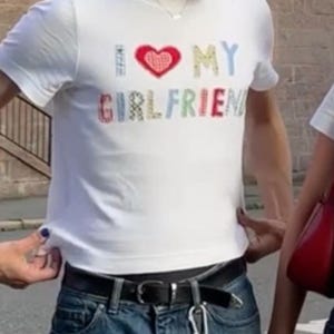 May include: A white short-sleeved t-shirt featuring the text I HEART MY GIRLFRIEND printed in multicolored, patterned letters across the chest, with a red heart symbol replacing the word heart. The shirt has a crew neckline and a straight hem.