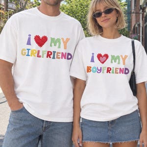 May include: Two white t-shirts featuring colorful, patterned, stitched-style lettering. One shirt displays the text I HEART MY GIRLFRIEND with a red heart, and the other displays I HEART MY BOYFRIEND with a red and white checkered heart.