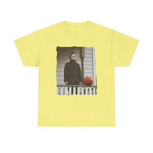 May include: A yellow t-shirt featuring a graphic print of a man wearing a white mask and holding a jack-o'-lantern. The man is standing on a porch with a white railing.