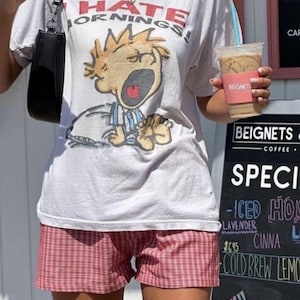 Op de afbeelding: Een wit T-shirt met een afbeelding van een cartoonfiguur dat "Ik haat ochtenden!" zegt. De persoon draagt een rood-wit geruite broek en houdt een kopje ijskoffie vast.