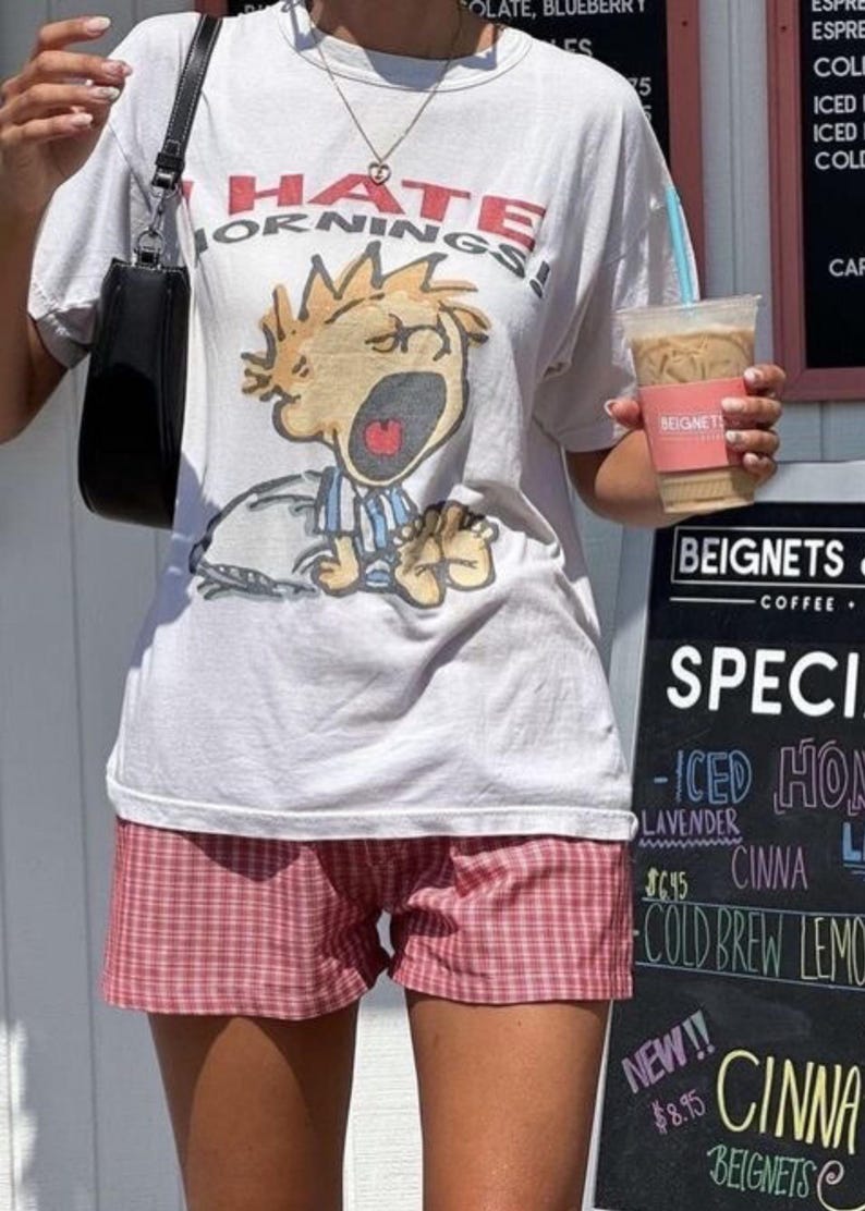 Op de afbeelding: Een wit T-shirt met een afbeelding van een cartoonfiguur dat "Ik haat ochtenden!" zegt. De persoon draagt een rood-wit geruite broek en houdt een kopje ijskoffie vast.