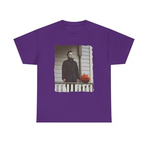 May include: Purple t-shirt featuring a graphic of Michael Myers from the Halloween film series standing on a porch with a jack-o'-lantern.