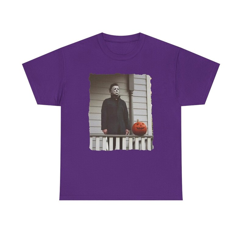 May include: Purple t-shirt featuring a graphic of Michael Myers from the Halloween film series standing on a porch with a jack-o'-lantern.
