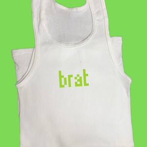 May include: White ribbed tank top with the word "brat" in green pixelated font. The top has a scoop neck and a sleeveless design. The background is a solid lime green color.