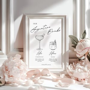 May include: A framed sign with the text "Our Signature Drinks" in elegant script. The sign features illustrations of two cocktails, one labeled "His" and the other "Hers", with their ingredients listed below. The frame is white, and the background is a soft white.