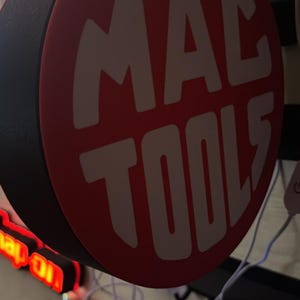 May include: A circular sign, red with white text reading "MAC TOOLS". The sign is mounted on a wall. Other signs and electrical cords are visible in the background.