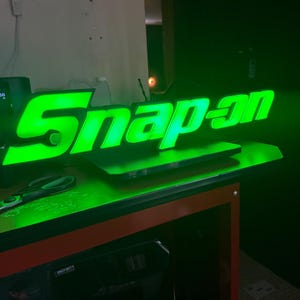 May include: A glowing green "Snap-on" sign, illuminated against a dark background. The sign is placed on a red and black surface, with a laptop and other items visible below. The letters are bold and modern.