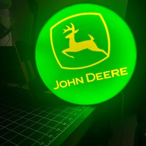 May include: A circular, illuminated sign featuring the bright green and yellow John Deere logo. The design includes a yellow leaping deer silhouette inside a yellow-bordered shape, with the text JOHN DEERE printed in yellow below the deer.