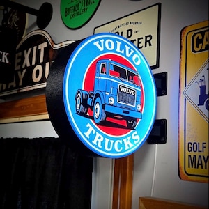 May include: A circular, illuminated wall-mounted sign featuring a blue Volvo truck graphic centered on a red circle. The sign has a black frame and the text VOLVO TRUCKS in white lettering against a blue background. A white power cord extends downward.