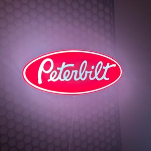 May include: A red, oval-shaped Peterbilt sign with white lettering and a white border. The sign is illuminated and mounted on a wall with a honeycomb pattern.