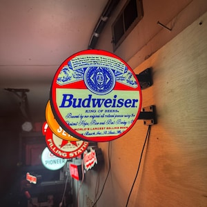 Puede incluir: Un letrero circular e iluminado de Budweiser montado en una pared de madera. El letrero presenta el logotipo de Budweiser en rojo, blanco y azul con el texto "Budweiser King of Beers" y "Brewed by our original all natural process" en azul.