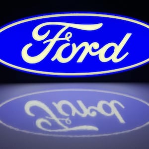 May include: A blue oval sign with the word "Ford" in white script. The sign is illuminated, and its reflection is visible below. The background is dark, emphasizing the bright logo.