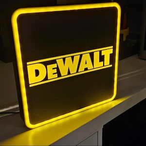 Op de afbeelding: Verlicht vierkant bord met het woord "DEWALT" in gele letters op een zwarte achtergrond. Het bord heeft een gele rand die oplicht. Het bord is aangesloten en staat op een wit oppervlak.