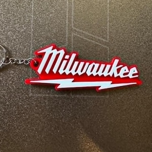 May include: A red and white Milwaukee keychain with a silver key ring and chain. The keychain features the word "Milwaukee" in white, with a red outline and a white lightning bolt design.
