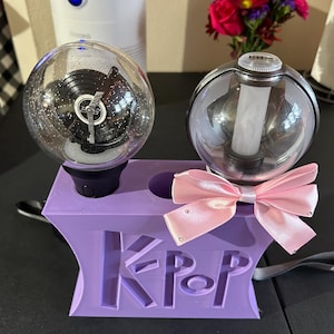 May include: Two clear, spherical light sticks with internal designs, resting in a purple holder with the letters "KPop" cut out. A pink bow with rhinestones is attached to the holder. The light sticks are on a black surface.