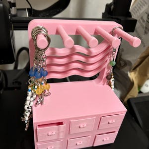 May include: A pink, 3D-printed miniature dresser with six drawers and a curved back. It has five pegs on top for hanging items, and several keychains with beads and charms are hanging from the pegs.