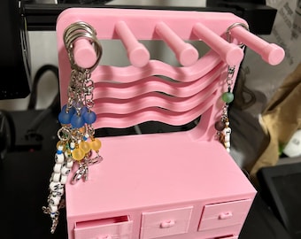 以下が含まれることがあります： ピンク色の3Dプリントされたミニチュアのドレッサー。6つの引き出しと湾曲した背面があります。上部には5つのペグがあり、アイテムを吊るすことができ、ビーズやチャームが付いたいくつかのキーチェーンがペグから吊り下げられています。