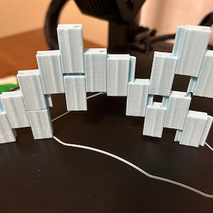 May include: A light blue, 3D-printed articulated sculpture composed of rectangular blocks. The blocks are connected in a zig-zag pattern, creating a flexible structure. The sculpture is resting on a black surface, with a white wire extending from it.