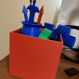 May include: A red square container holding various 3D-printed objects. Inside, a blue pot with blue arrows and a red sword is visible. Additional blue and green objects are also present, suggesting a storage or organizational theme.
