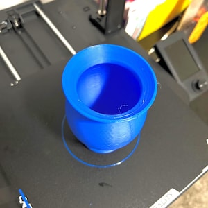 May include: A vibrant blue, 3D-printed cylindrical object with a broad, flat rim. The object rests on a black surface, probably a 3D printer bed. The object has a smooth texture and a rounded interior.