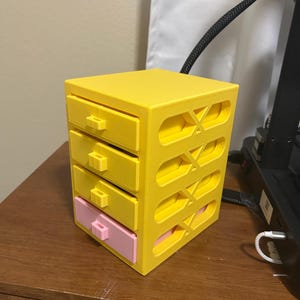 May include: A yellow 3D-printed storage box with four drawers, one pink and three yellow. The box has an open design with decorative cutouts on the sides. The drawers have small, rectangular handles.