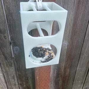 Puede incluir: Un comedero para pájaros rectangular de color blanco roto con una superficie texturizada y dos aberturas circulares en la parte frontal. El comedero contiene un pequeño montón de semillas de girasol negras y granos mezclados en la bandeja.