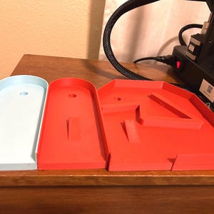 May include: Three 3D-printed miniature golf course elements. One is light blue, and the other two are red. The red pieces have angled walls and obstacles. The items are on a wooden surface.