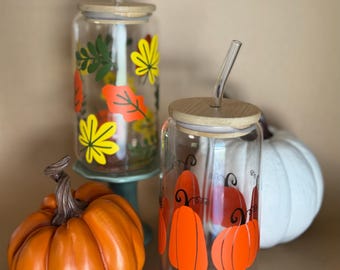 May include: Two glass tumblers with wooden lids and reusable straws. One tumbler has a fall leaf design in yellow, orange, and green. The other tumbler has a pumpkin design in orange. Both tumblers are sitting on a table with two pumpkins.