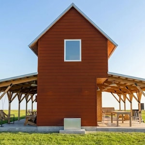Può includere: Un edificio in stile fienile a due piani, marrone-rossastro, con tetto a capanna e una piccola finestra. L'edificio ha portici coperti su entrambi i lati, sostenuti da travi di legno. Sono visibili un'altalena e posti a sedere.