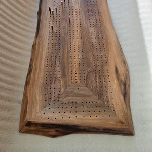 Può includere: Un tabellone di cribbage in legno fatto a mano con un bordo naturale. Il tabellone è realizzato in legno marrone scuro e presenta una serie di piccoli fori per i pioli. Sono inseriti diversi pioli metallici di vari colori.