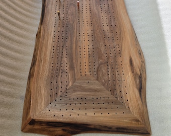 May include: A handcrafted wooden cribbage board with a live edge. The board is made of dark brown wood and features a series of small holes for pegs. Several metal pegs in various colours are inserted into the board.