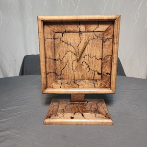Op de afbeelding: Een houten klok met een donkerbruine vlek en een goudkleurig gezicht. De klok heeft een vierkant gezicht en een standaard. Het hout heeft een uniek gebarsten patroon.