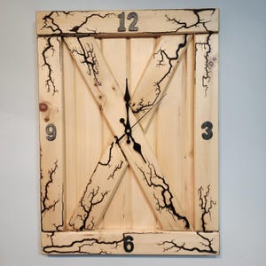 May include: A wooden clock with a barn door design. The clock face has black hands and black Roman numeral markings. The wood has a light brown stain and black lightning bolt patterns.
