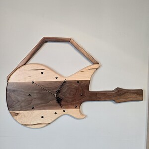 Könnte beinhalten: Eine Wanduhr in Gitarrenform aus Holz mit einem hell- und dunkelbraunen Streifendesign. Die Uhr hat schwarze Zeiger und schwarze Punkt-Stundenmarkierungen. Über der Gitarre befindet sich ein Holzrahmen.